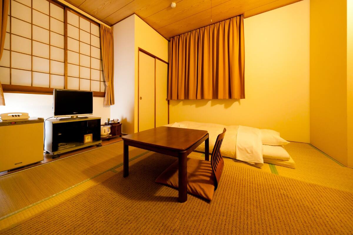 Una habitación japonesa tradicional con suelo de tatami, una mesa de madera baja, un televisor sobre un mueble y una cama con ropa de cama blanca.