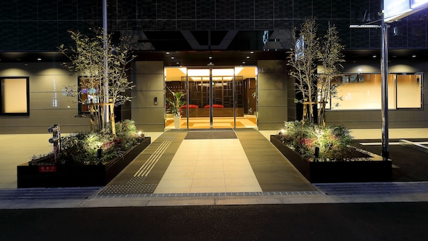 A hotel entrance with a modern design, featuring a glass door, a tiled pathway, and landscaped planters.