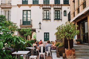 Un café en plein air, avec des tables et des chaises, entouré d’immeubles aux volets verts et à la façade blanche.