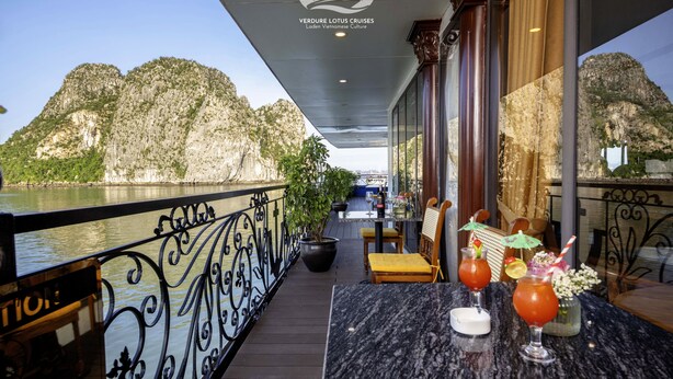 Un balcón con vista a un río y montañas, una mesa con bebidas, y un letrero de Verdure Lotus Cruises.