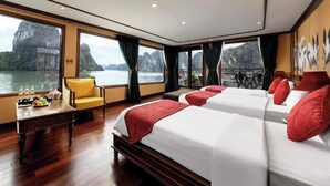 Habitación de hotel con dos camas, un escritorio de madera con una silla, una mesa con botellas y fruta, y ventanas amplias con vistas al agua y montañas.