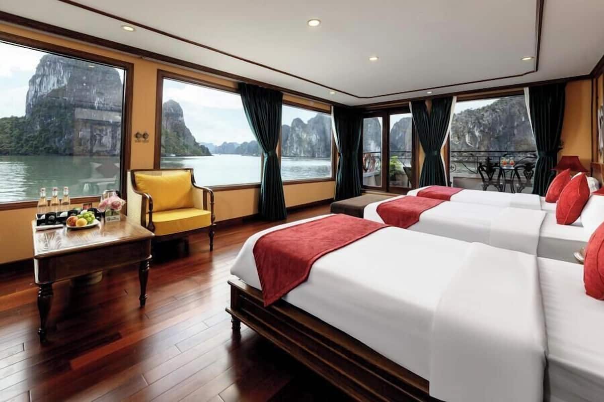 Habitación de hotel con dos camas, un escritorio de madera con una silla, una mesa con botellas y fruta, y ventanas amplias con vistas al agua y montañas.