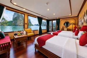 A room with a large window offering a view of a body of water and cliffs, a wooden floor, a desk with a chair, and two beds with red bedding. - Verdure Lotus Grandeur Cruise on Ha Long Bay (Tuan Chau)