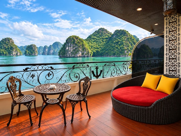A balcony with a view of a turquoise sea and green islands, furnished with wicker chairs and a red sofa. - Verdure Lotus Grandeur Cruise on Ha Long Bay (Tuan Chau)