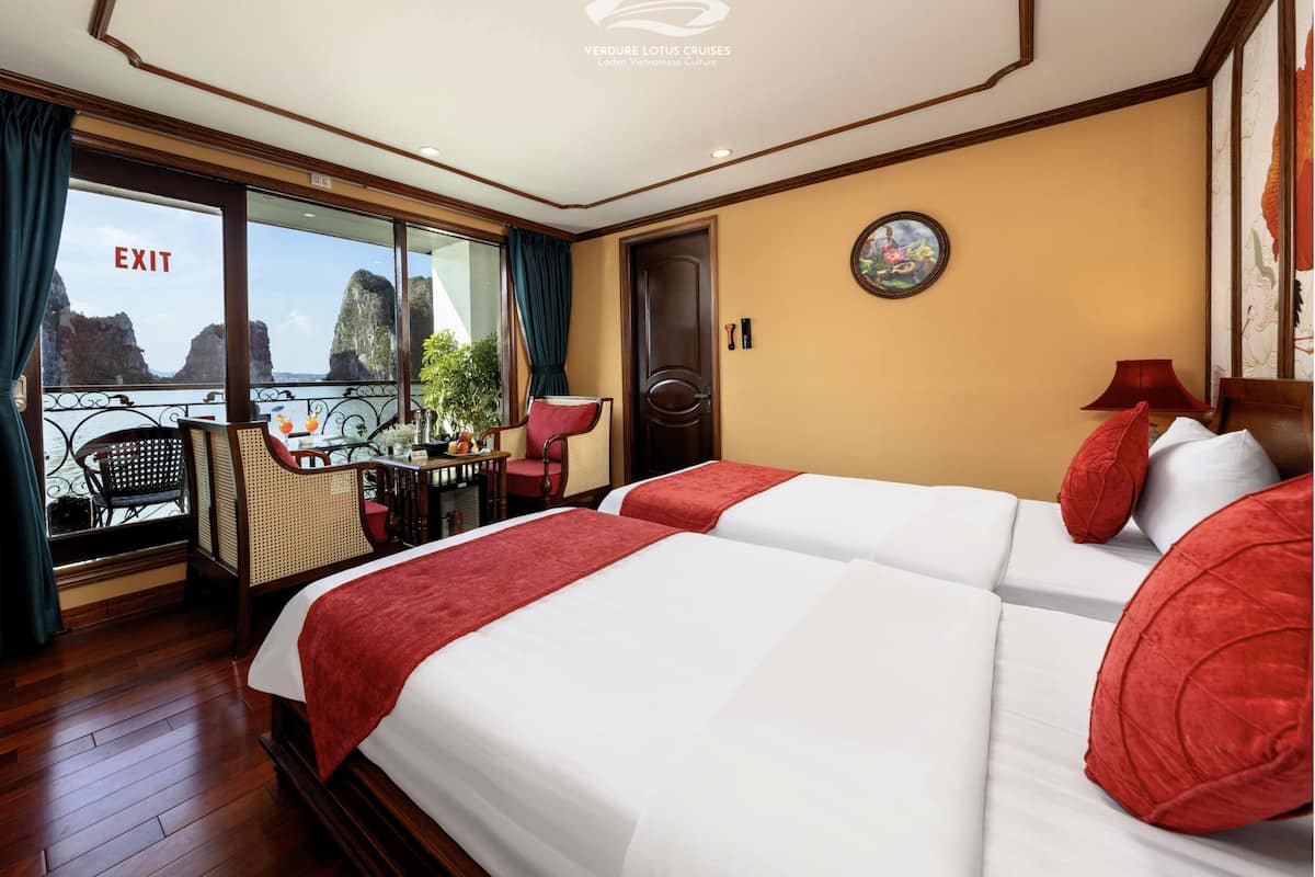 Habitación de hotel con balcón, cama con almohadas rojas, una mesa con una planta y un cuadro en la pared.