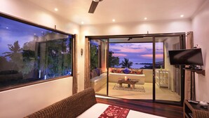 A modern hotel room with a large window, a ceiling fan, a flat-screen TV, a sofa, and a balcony with a view of the sea. - Villa Umbrella (Senggigi)