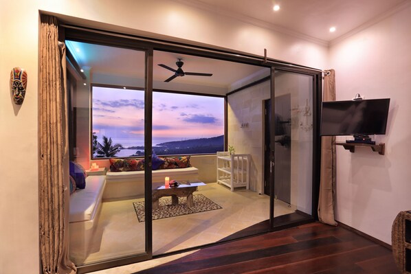 A modern living room with a glass door leading to a balcony, a ceiling fan, and a mounted TV. - Villa Umbrella (Senggigi)