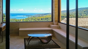 A room with a large window offering a view of a coastal landscape, a small table, and a bench. - Villa Umbrella (Senggigi)