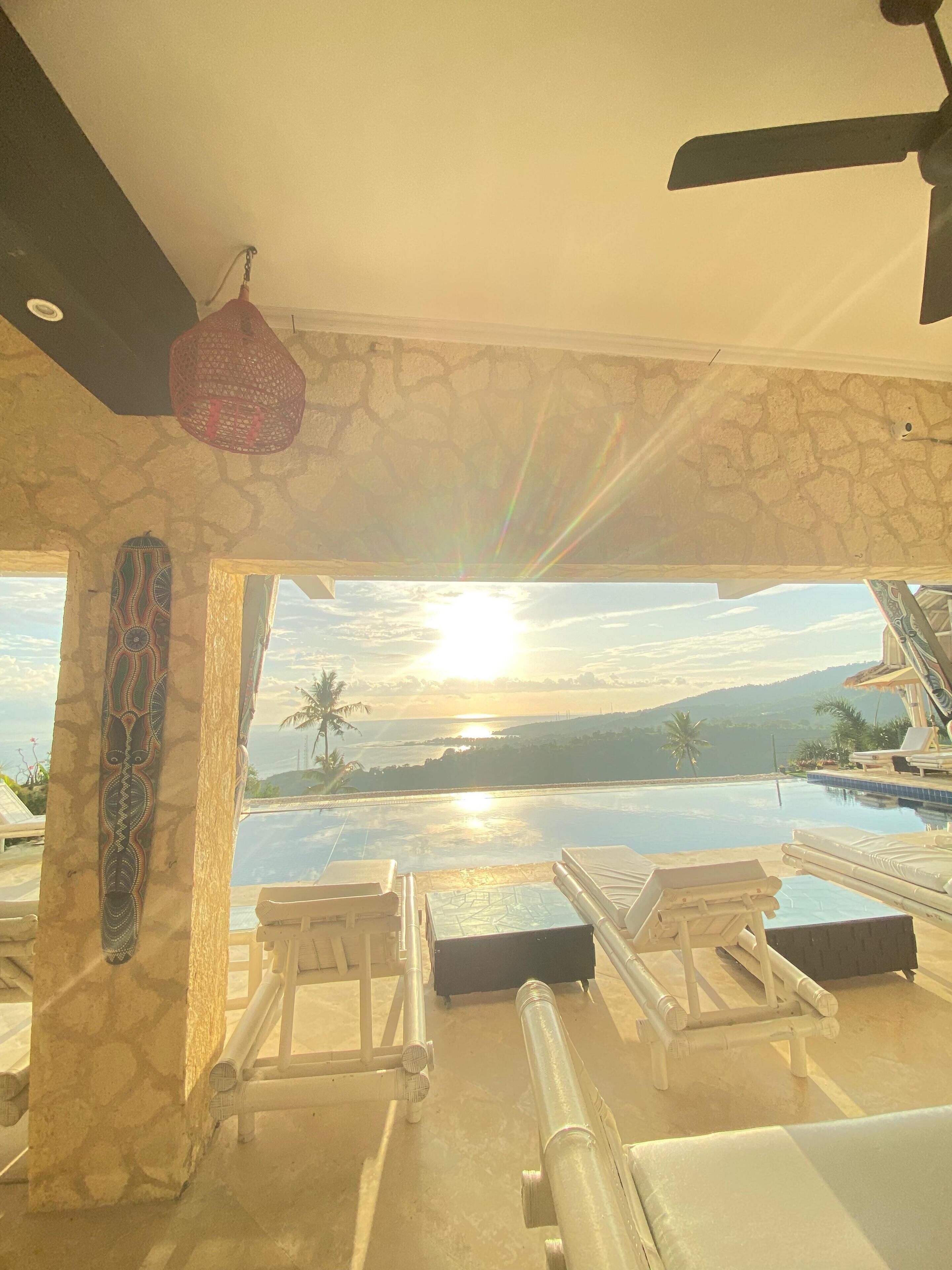 A sunlit room with a view of a pool and sunset, stone walls, and lounge chairs.