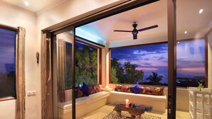 A modern living room with a glass door leading to a balcony, a ceiling fan, and a seating area with cushions. - Villa Umbrella (Senggigi)