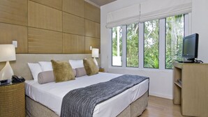 A modern hotel room with a large bed, a television, and a window with a view of greenery. - Beach Club Port Douglas 3 Bedroom Luxury Apartment (Port Douglas)