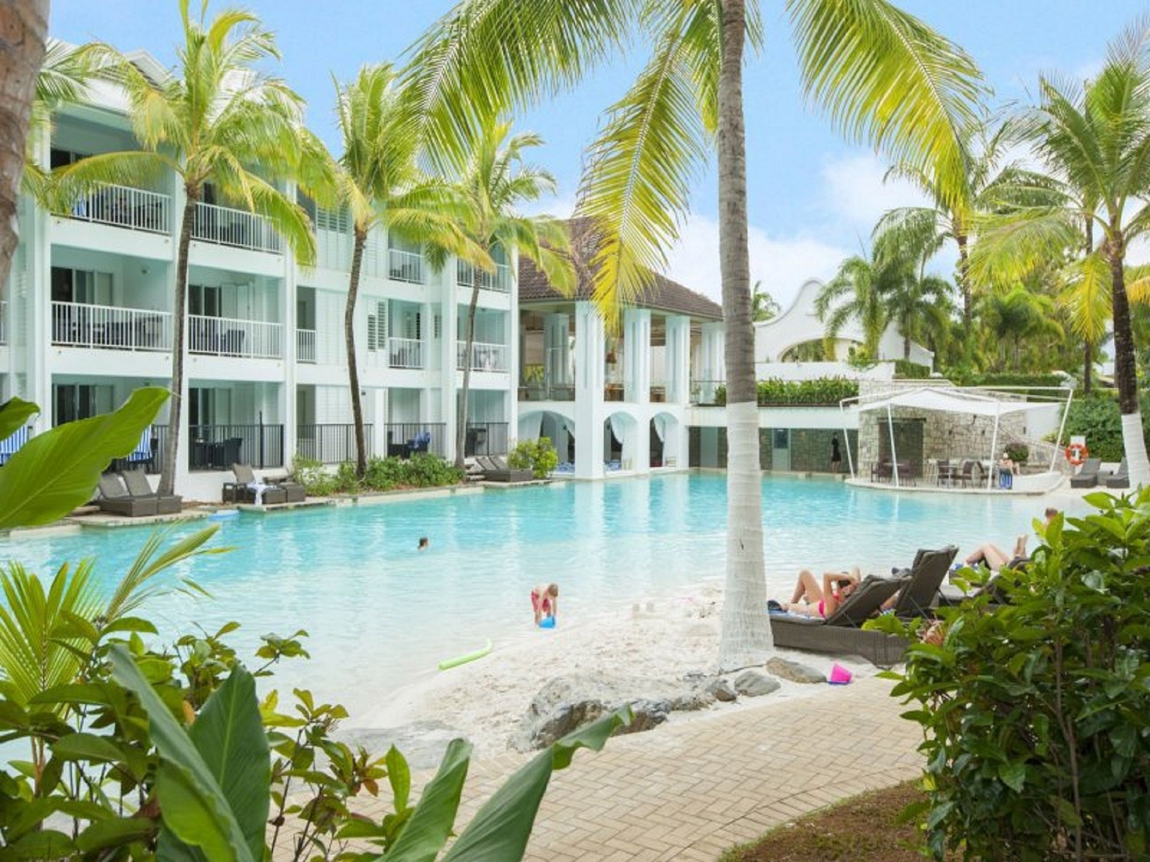 A resort with a pool surrounded by palm trees and a multi-story building with balconies.