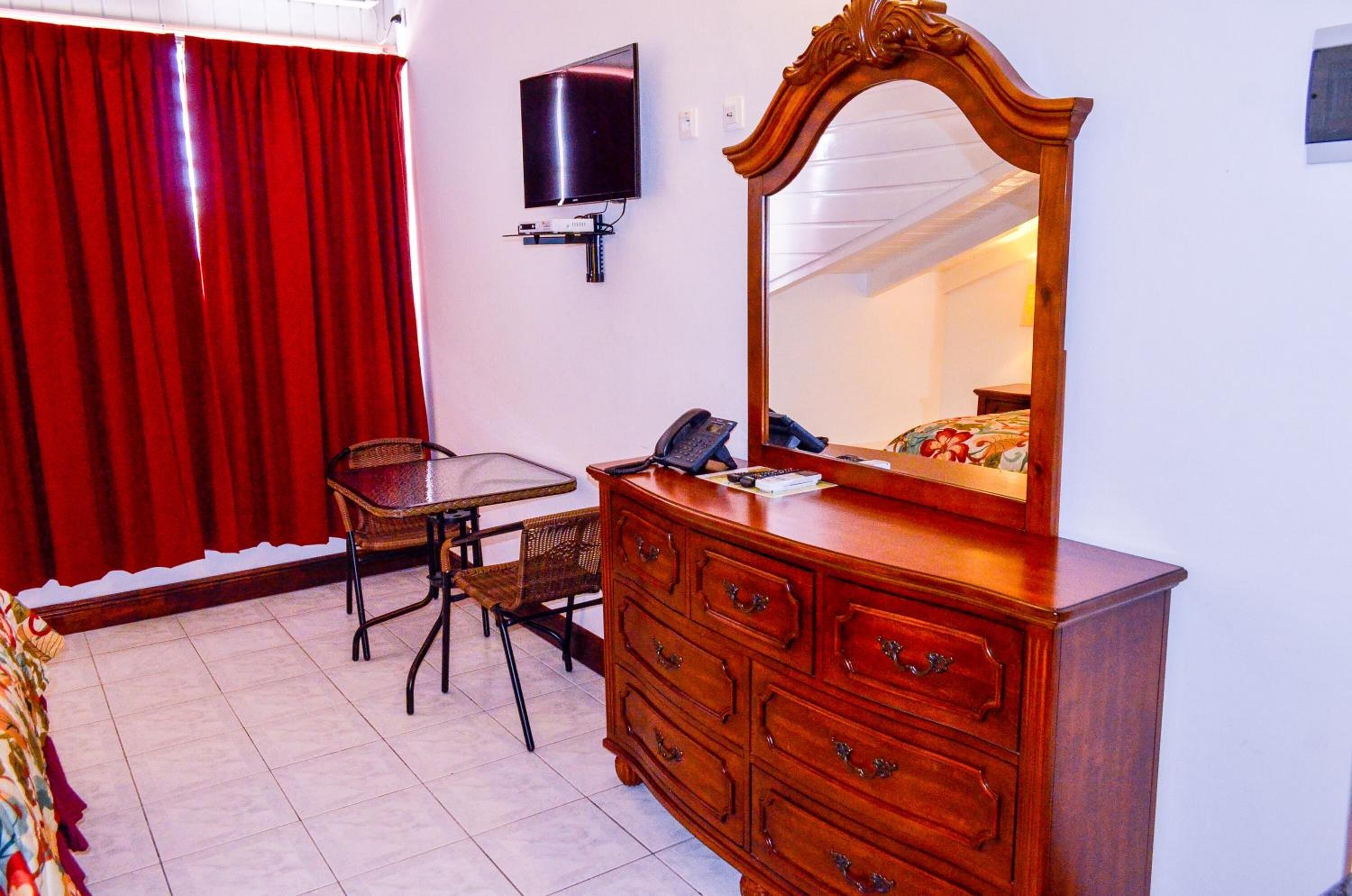 A bedroom with a wooden dresser, a mirror, a television mounted on the wall, a small table with chairs, and a red curtain.