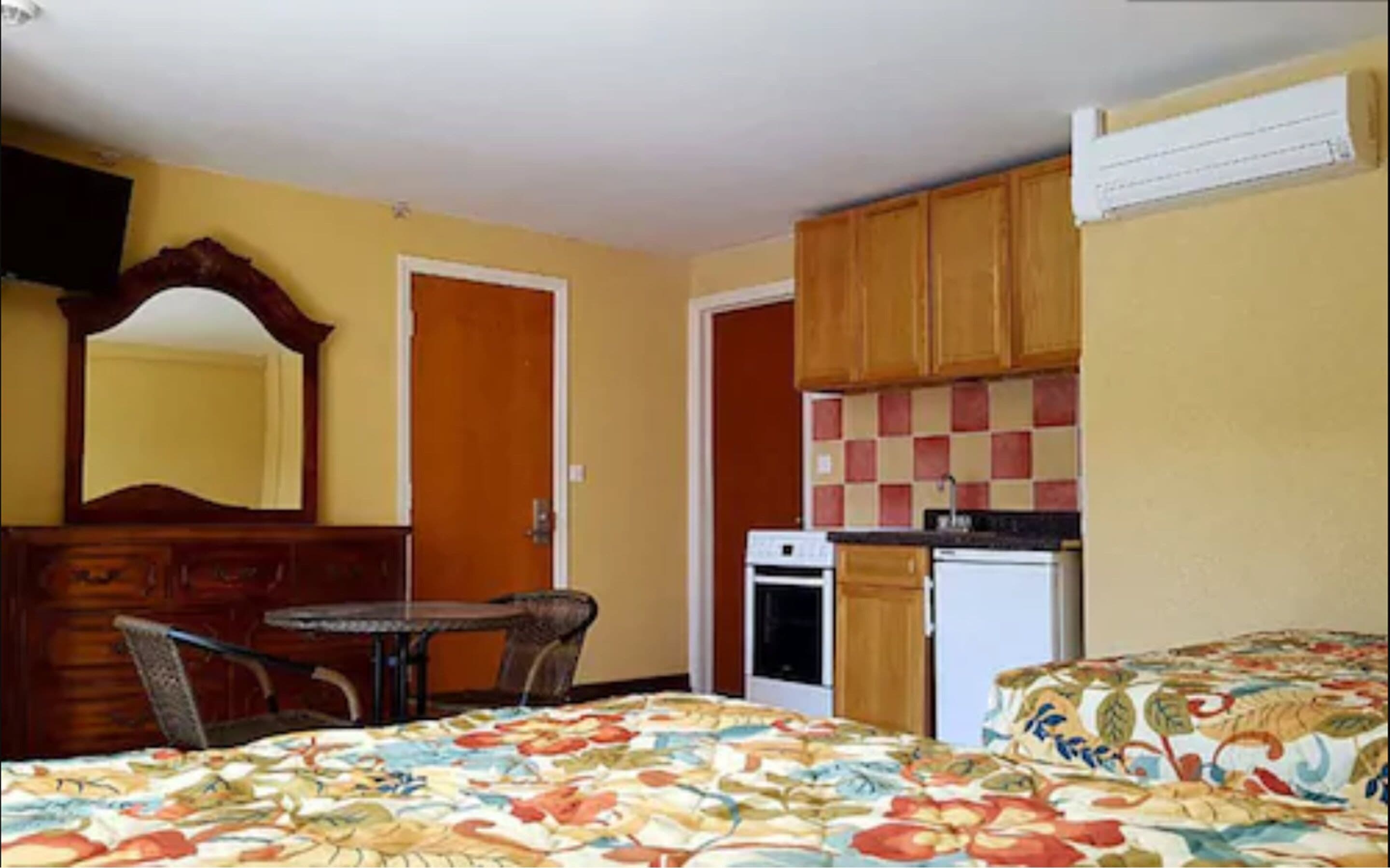 A bedroom with a bed, a chair, a desk, a mirror, and a kitchenette with a stove and refrigerator.