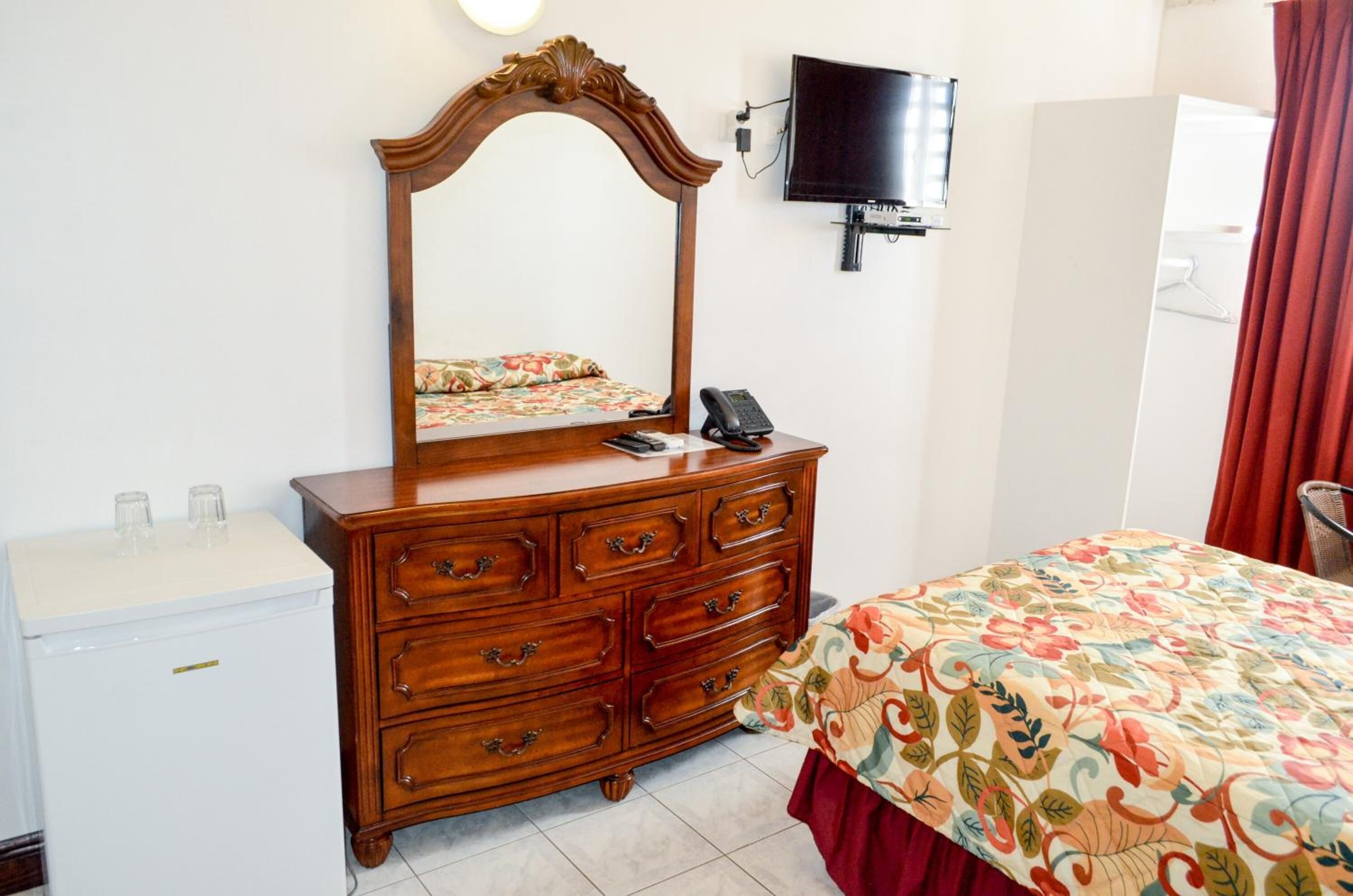 Una habitación de hotel con un aparador de madera, una cama con colcha estampada, un televisor montado en la pared y un mini refrigerador blanco.