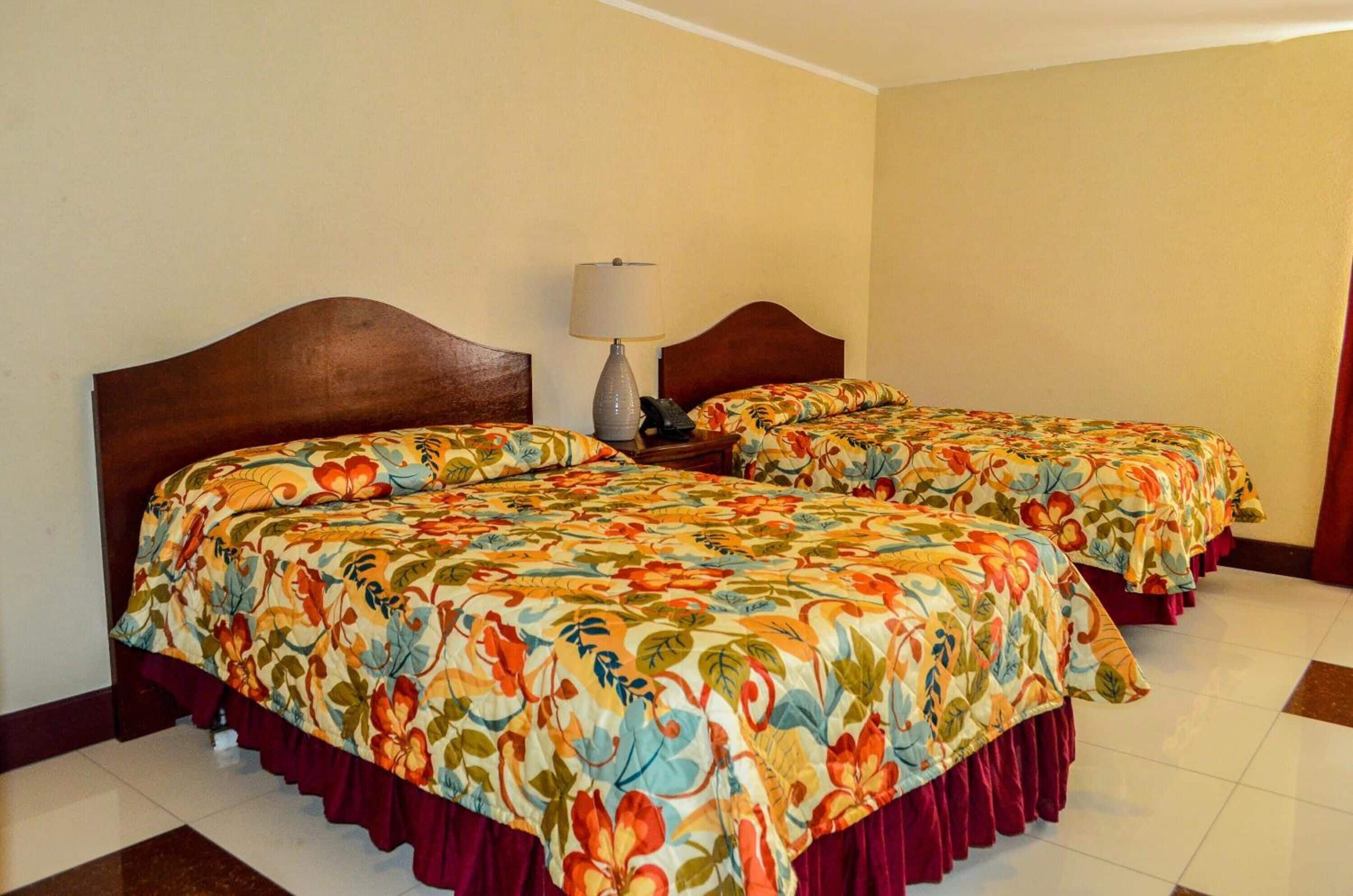 Habitación de hotel con dos camas, cada una con ropa de cama floral y cabeceros de madera. Hay una mesita de noche con una lámpara y un teléfono entre las camas.