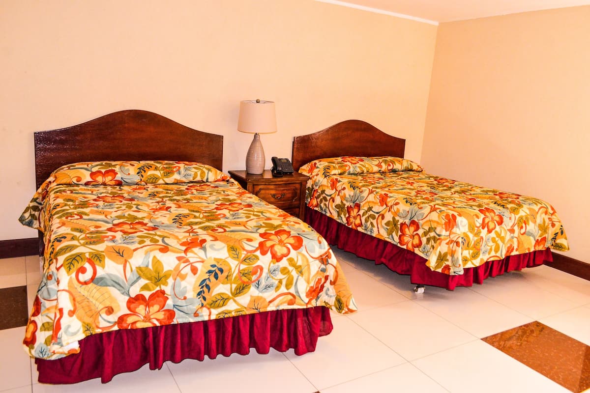 Two single beds with floral bedding, a nightstand with a lamp and phone, and a tiled floor.