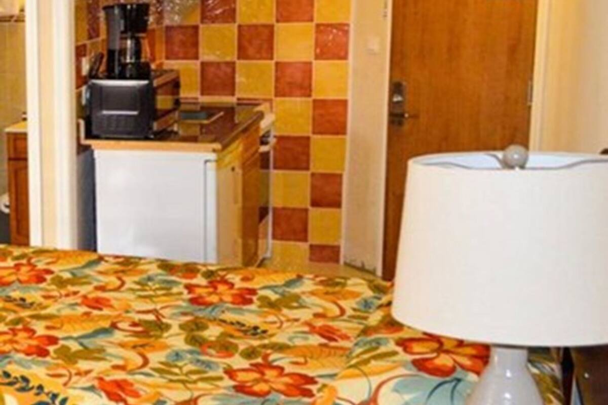 A small, cozy room with a bed, a bedside lamp, a wooden cabinet, and a kitchenette.