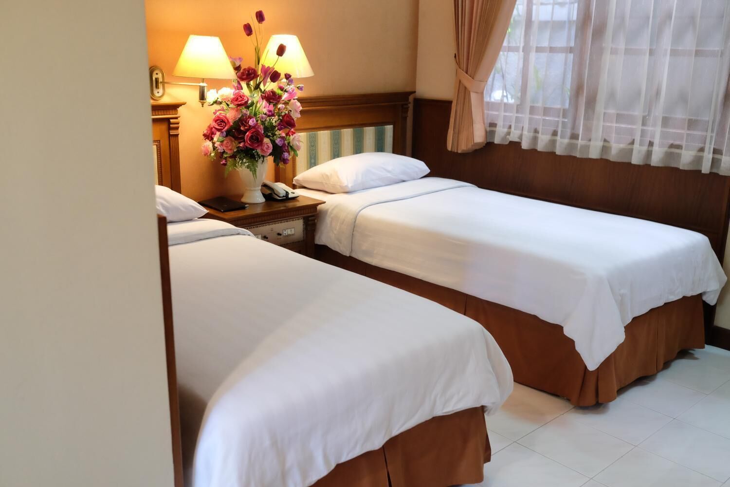 Una habitación de hotel con dos camas individuales, una mesita de noche con lámpara y teléfono, y un arreglo floral.