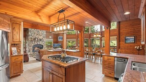 A spacious kitchen with a central island, stone fireplace, wooden ceiling, and large windows. - Donner Lake Retreat | Queen Suite, Games, Near Lake, Golf & Trails (Truckee)
