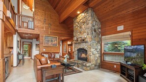 A spacious living room with a stone fireplace, wooden ceiling, and a large TV. - Donner Lake Retreat | Queen Suite, Games, Near Lake, Golf & Trails (Truckee)