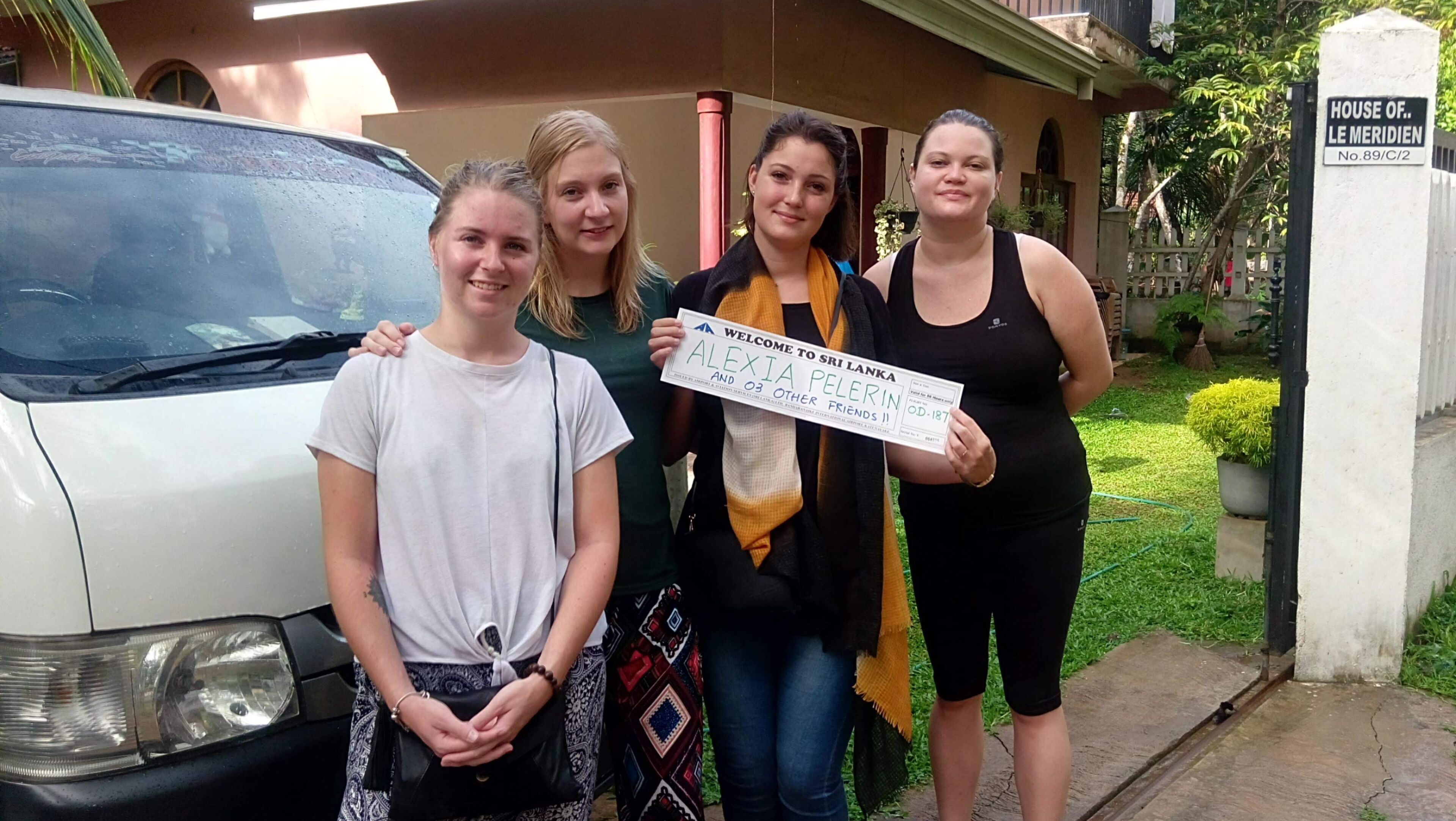 Four people standing in front of a house with a sign that reads "HOUSE OF.. LE MERIDIEN No.89/C/2".