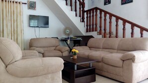 Una sala de estar con dos sofás beige, una pequeña mesa con un florero, un televisor, un ventilador y una escalera con barandilla de madera.