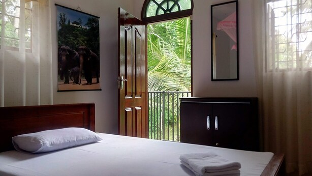 Habitación de hotel con cama, cabecera de madera, cuadro enmarcado con elefantes, ventana con vista a palmeras y un armario de color oscuro.