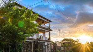 Una casa de dos plantas con balcón, rodeada de follaje y una entrada protegida por una reja.