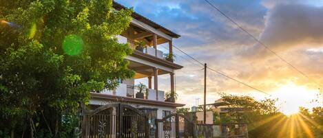 A two-story house with a balcony, surrounded by greenery, and a gated entrance.
