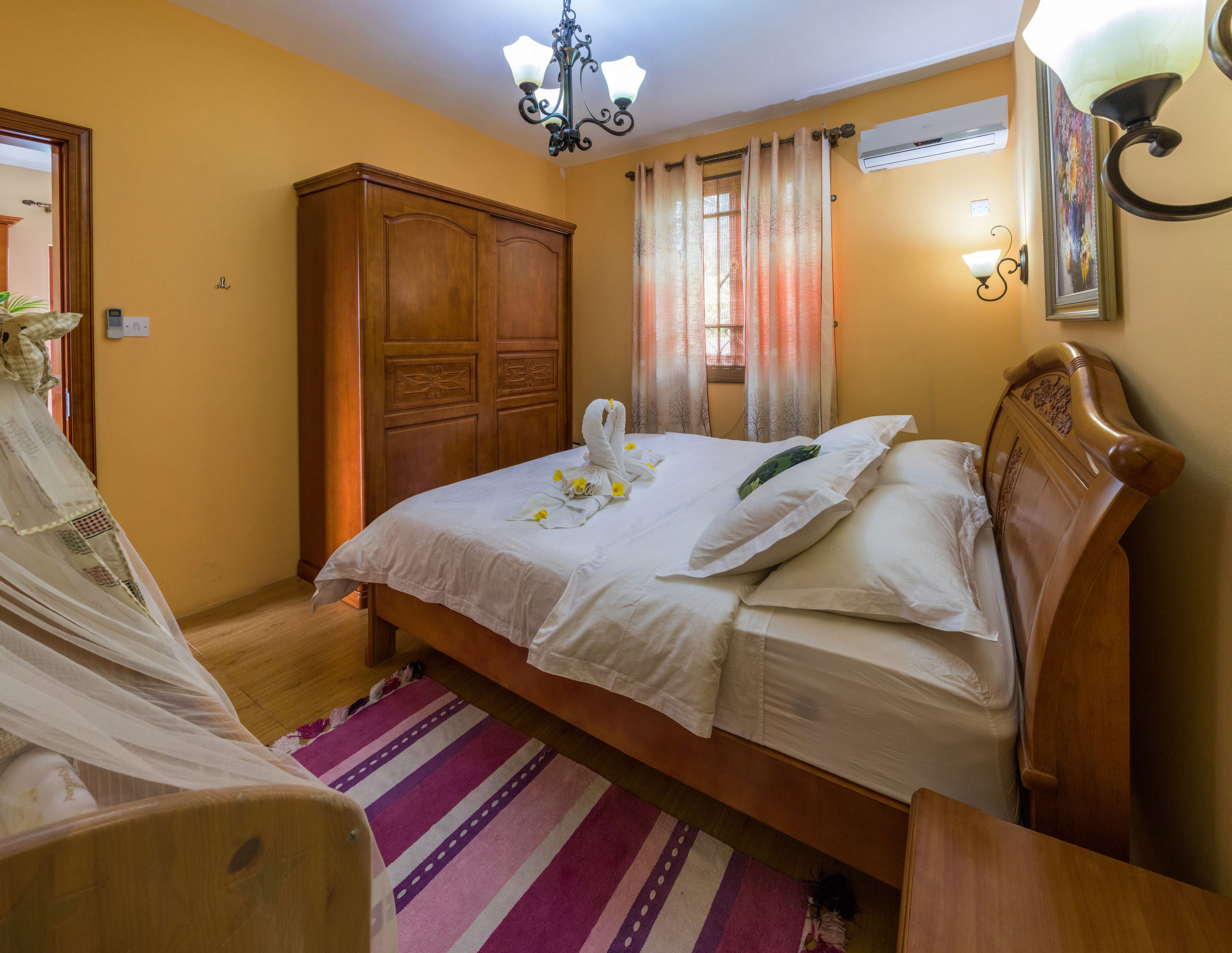 A neatly made bed with a swan-shaped pillow and a bouquet of flowers, a wooden wardrobe, a striped rug, and a chandelier.