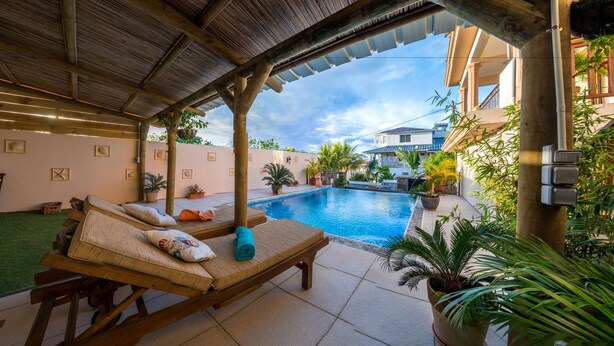 Zona junto a la piscina con sillones de descanso, un área de estar cubierta y vista a un edificio residencial al fondo.