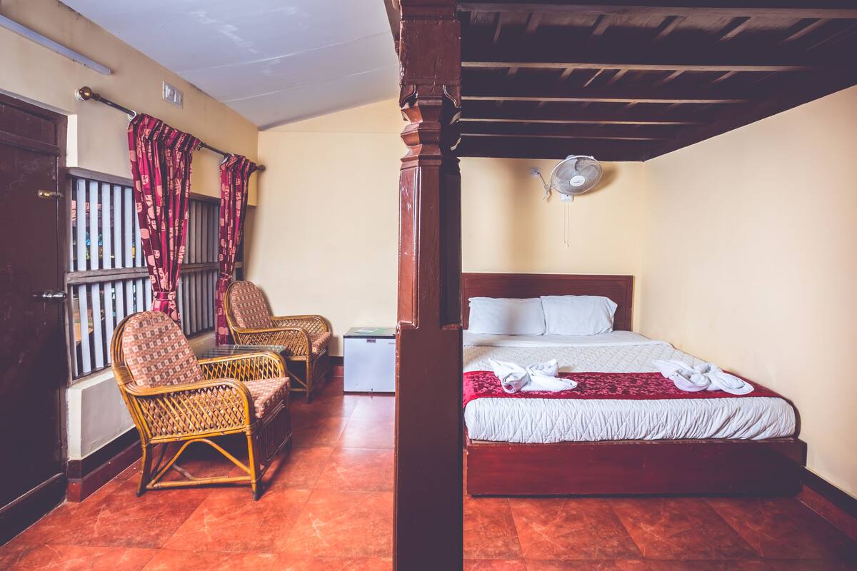 Une chambre d’hôtel avec un lit en bois, deux fauteuils en osier, une petite table et une fenêtre avec des rideaux.
