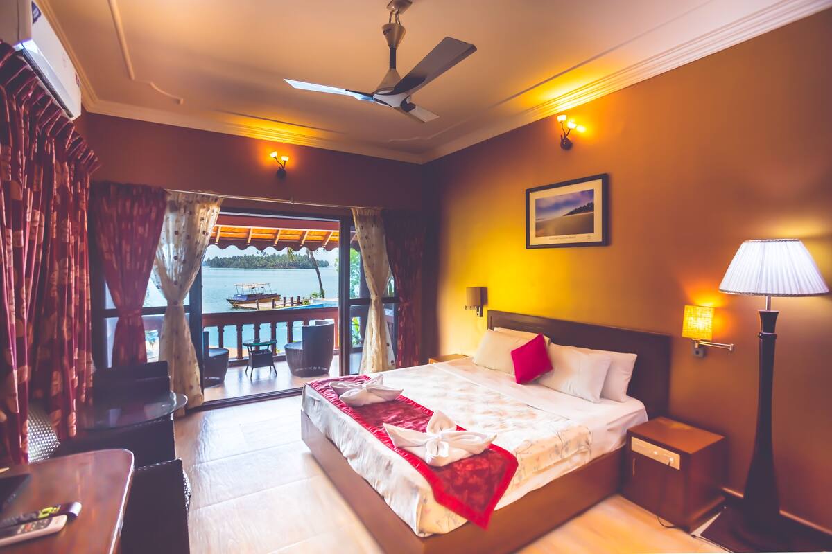 Une chambre d’hôtel avec un lit, une table de chevet, une lampe, un ventilateur de plafond et une vue sur une étendue d’eau, visible à travers de grandes fenêtres.