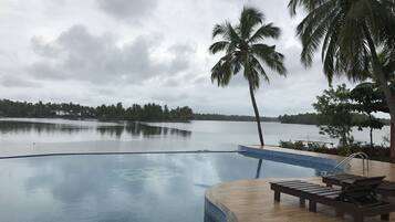 Piscina con cubierta de madera y sillas de descanso, rodeada de palmeras y vista a un lago tranquilo.