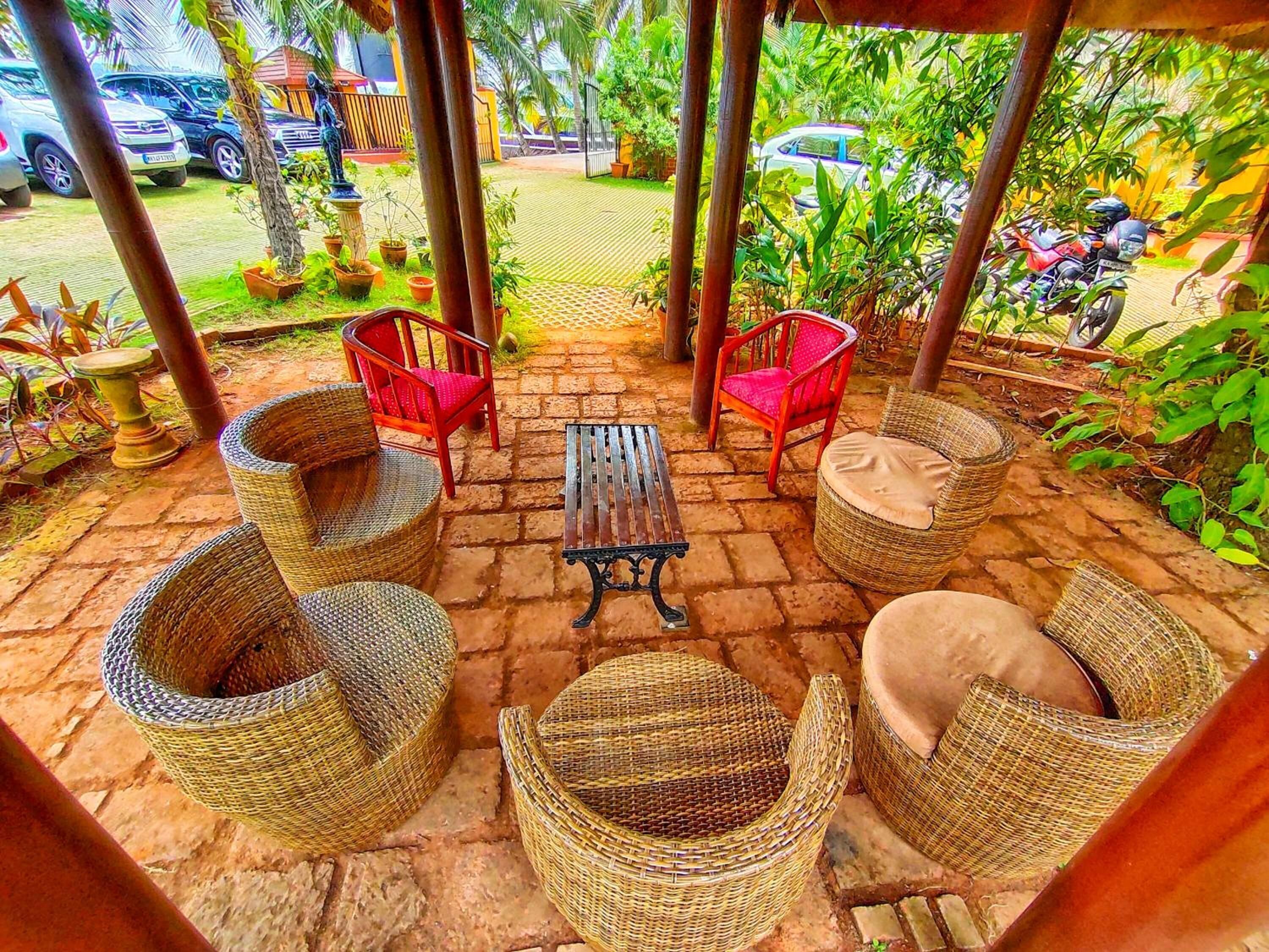 Un espace extérieur aménagé, abrité et doté de mobilier en osier, avec une table centrale et une vue sur un jardin et des voitures garées.