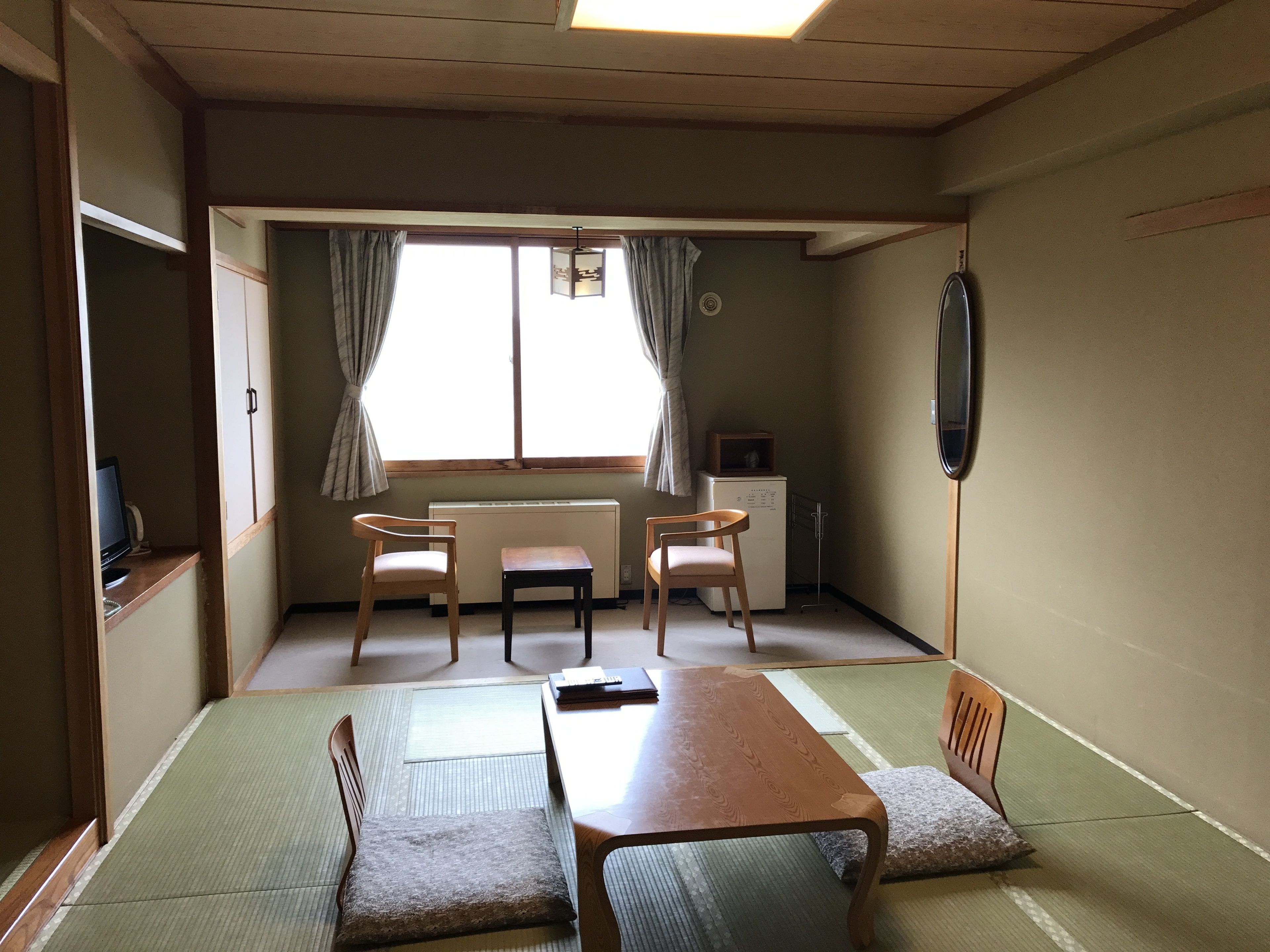 A traditional Japanese room with tatami flooring, a low table, two chairs, a small heater, and a television.