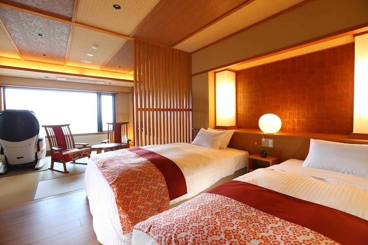 Habitación de hotel con dos camas, un área de estar con una silla y una mesa, y un ventanal que proporciona luz natural.