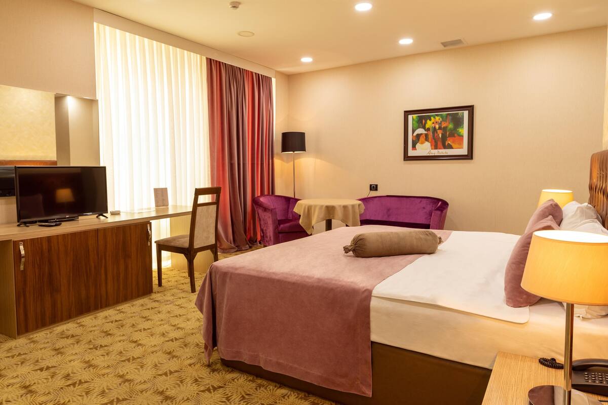 A hotel room with a bed, desk, TV, and a painting on the wall.