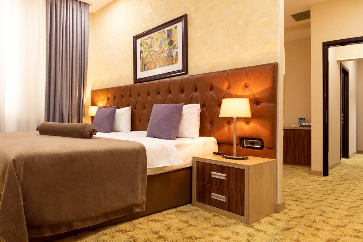 A hotel room with a large bed, a bedside table with a lamp, a nightstand, and a framed picture on the wall.
