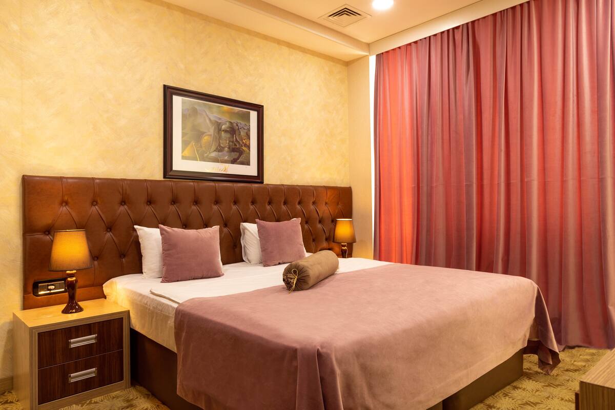 A hotel room with a large bed, a headboard, a nightstand with a lamp, a framed picture on the wall, and a window with curtains.