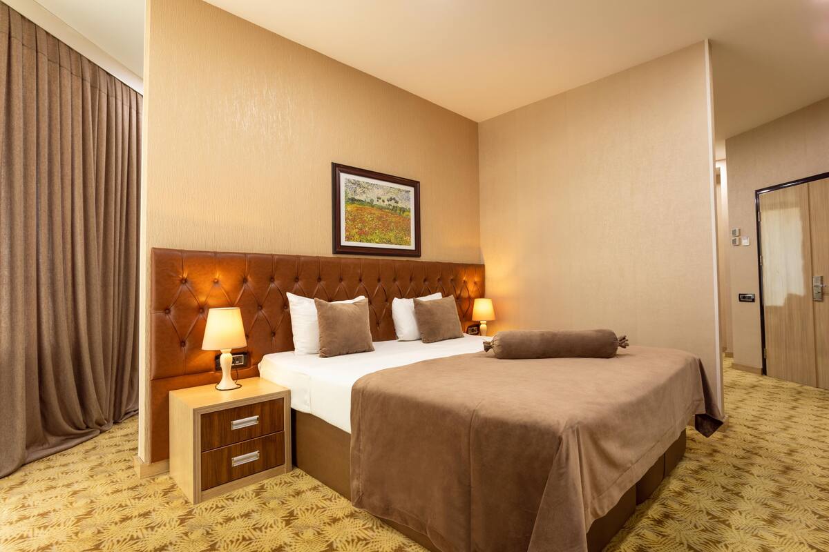 A hotel room with a large bed, a headboard, a nightstand, a lamp, a framed picture on the wall, and a door.