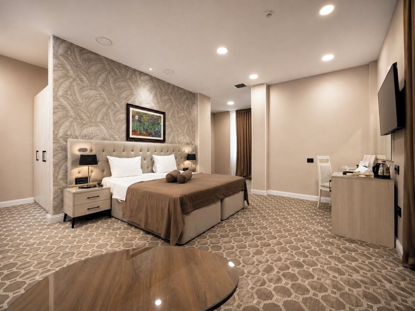 A hotel room with a large bed, a bedside table, a desk, a chair, a television, and a patterned carpet.
