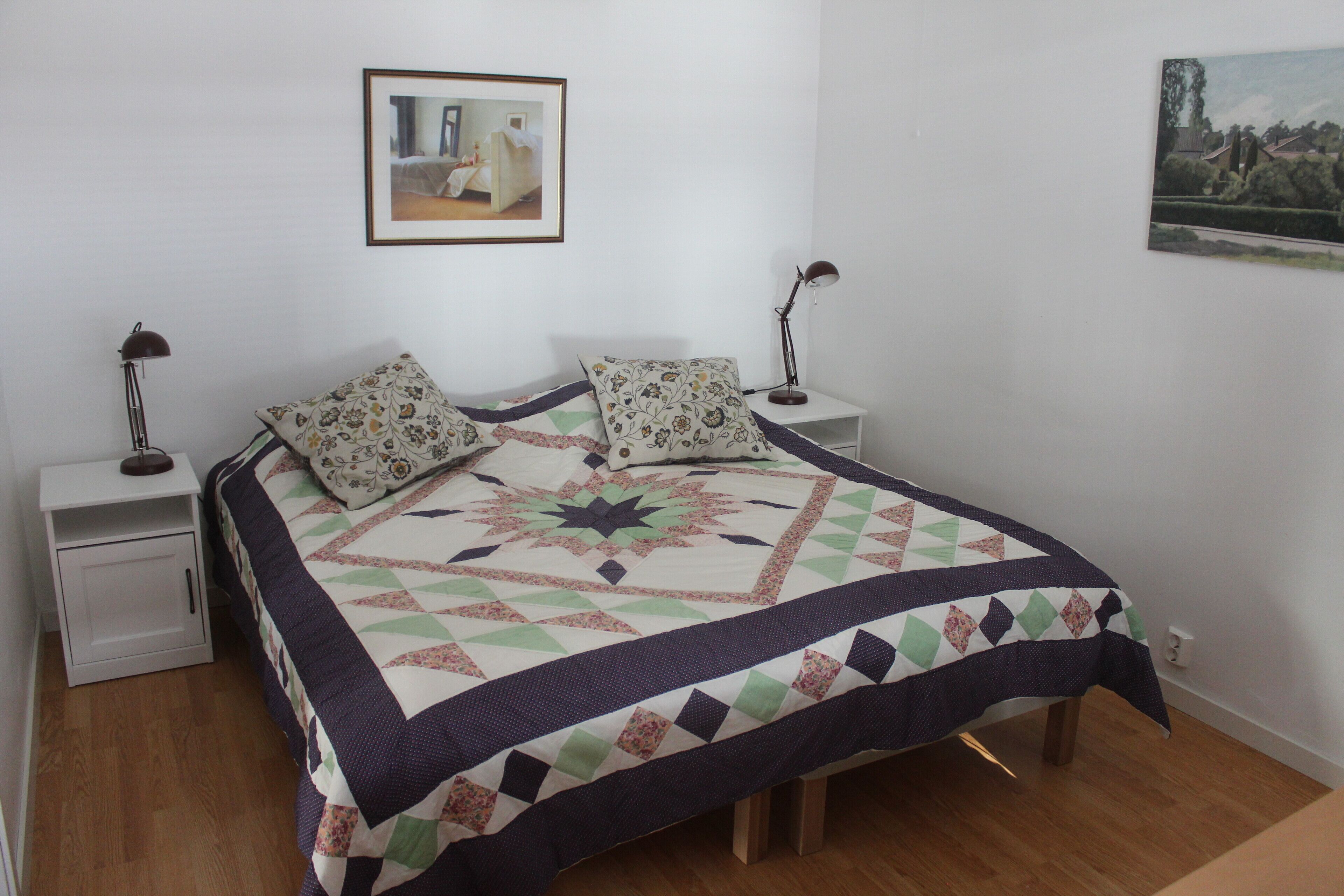 A neatly made bed with a patterned quilt, two bedside tables with lamps, and two framed pictures on the walls.