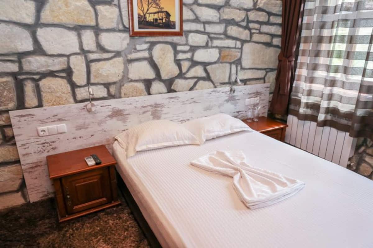 A hotel room with a stone wall, a wooden headboard, a neatly made bed with white linens, a bedside table with a remote control, and a framed picture on the wall.