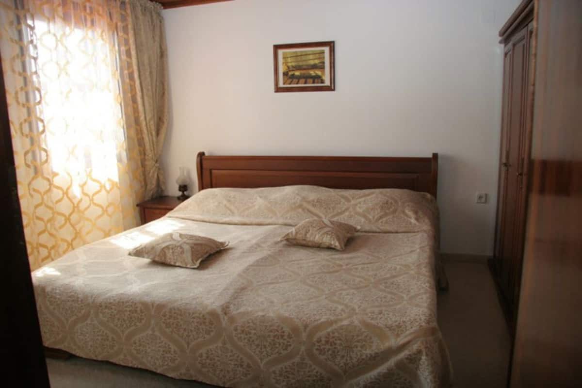 A bedroom with a wooden bed, patterned bedding, a nightstand, a wardrobe, and a framed picture on the wall.
