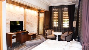A hotel room with a bed, a desk with a TV, two armchairs, a small table, and a window with curtains.