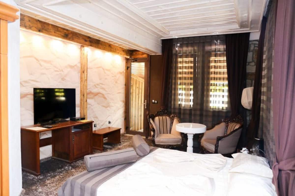 A hotel room with a bed, a desk with a TV, two armchairs, a small table, and a window with curtains.