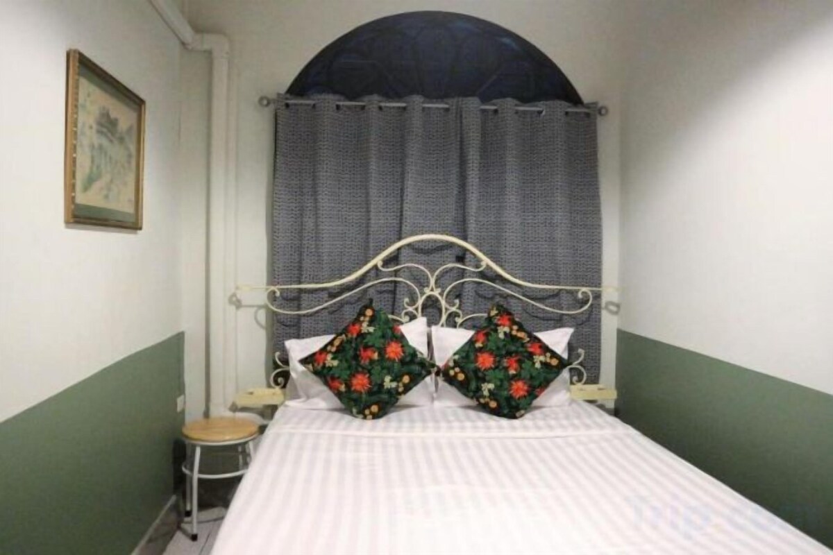 Una habitación con una cama, un taburete pequeño, un cuadro en la pared y una ventana con cortinas.