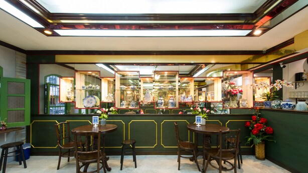 A restaurant with a green and yellow color scheme, featuring a counter with glass display cases and wooden chairs. - The Neighbors Hostel - Adults Only (Phuket)